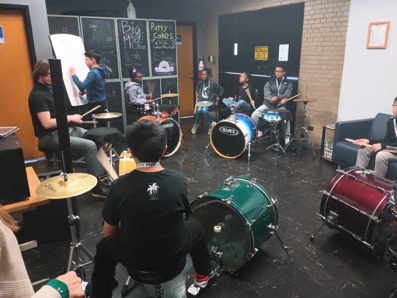 Students sit in a round circle, playing out rhythms on drumsets.