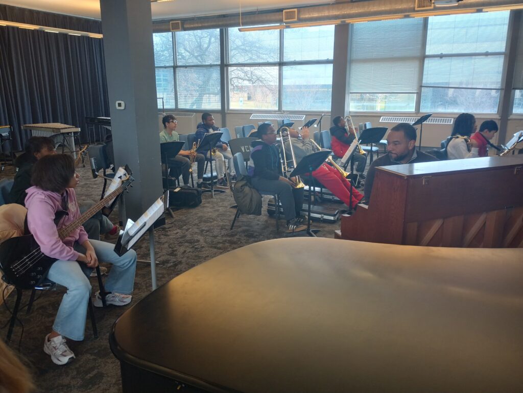 A jazz ensemble listens to their conductor. Visible is a baby grand piano, bass guitar, trombones, trumpets, and saxophones.