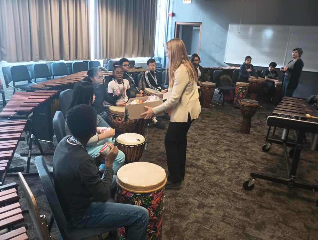 Dr. Schuman hands out a box of snacks to a percussion lesson. A few kids stare excitedly into the box.